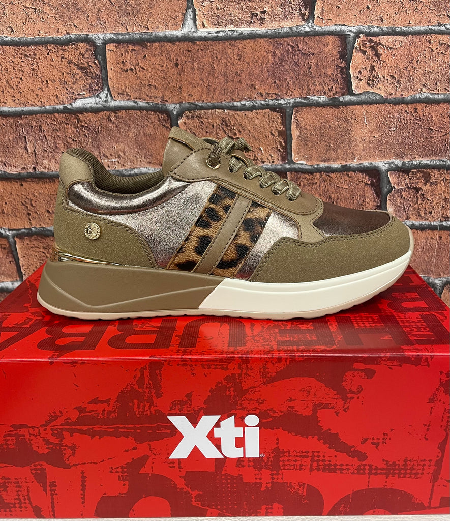 Gold sneaker with leopard print accents on a red Xti box against a brick wall.