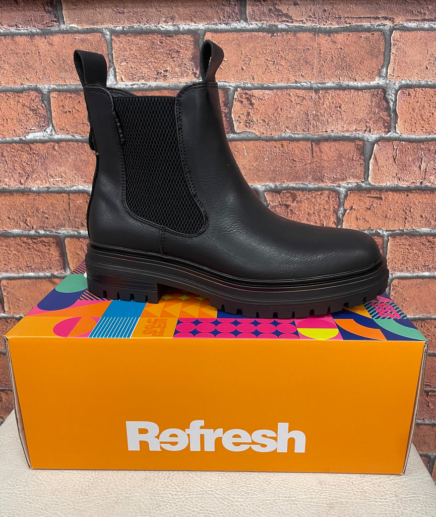 Black Chelsea boot on a colorful 'Refresh' box against a brick wall.