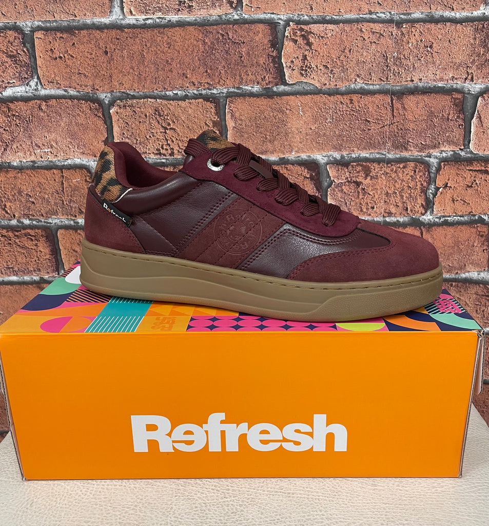 Maroon sneaker on a colorful box with 'Refresh' branding against a brick wall.